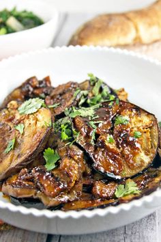 a white bowl filled with cooked eggplant and parsley on top of a wooden table