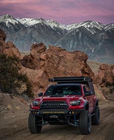 a red truck driving down a dirt road with mountains in the background