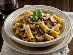 a white bowl filled with pasta and mushrooms on top of a table next to a glass of wine