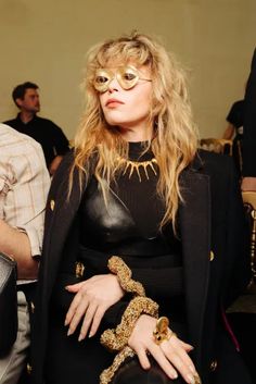 a woman sitting in a chair with her hands on her chest and wearing gold glasses