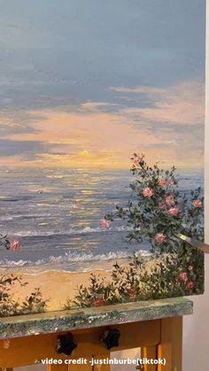 a painting is being displayed on a easel near the ocean and flowers are growing out of it