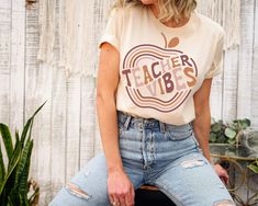 a woman sitting on top of a wooden bench next to a fence wearing a t - shirt that says teacher vibes