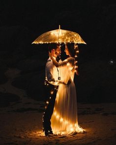 a bride and groom standing under an umbrella on the beach at night with fairy lights