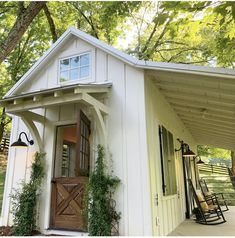 a small white shed with a porch and swing set on the front door is surrounded by greenery