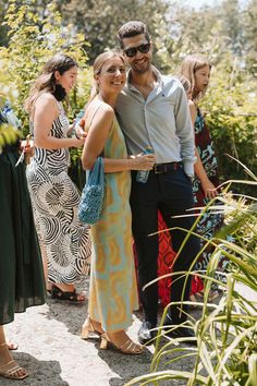 two people standing next to each other in front of some plants and trees with sunglasses on