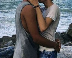 two people standing next to each other near the ocean and one is kissing the other