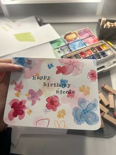 someone holding up a birthday card in front of some paintbrushes and watercolors