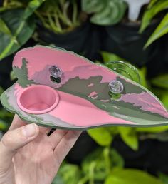 a hand holding a pink and green object in front of some potted plantes