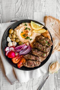 a black plate topped with meat, vegetables and bread next to a fork on a white table