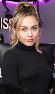 a woman standing in front of a purple background wearing a black turtle neck top and fur cuffs