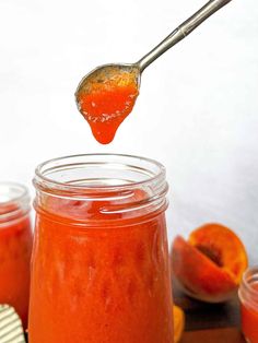 a spoon full of orange jam being lifted from a jar with two slices of peaches in the background