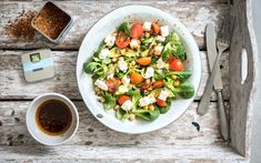 a white plate topped with salad next to a bowl of sauce and spoons on top of a wooden table