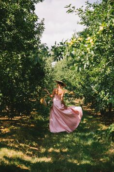 a woman in a pink dress and hat walking through an apple orchard with lots of trees