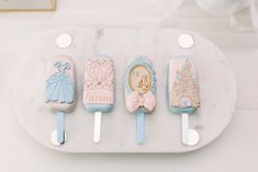 four ice cream pops decorated with princess themed icing on a marble platter next to a cupcake