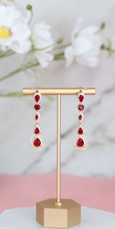 These earrings showcase a pear-shaped design, accentuated with ruby red cubic zirconia (CZ) stones that exude a rich, vibrant hue. They dangle gracefully, adding a pop of color and elegance to any ensemble, making them a perfect statement piece for special occasions or everyday wear. Length: 2 inches Width: 0.50 inches Closure: Bullet with Disc Backs Material: Brass with 18K Gold Plating with Rhodium Coating Lead Free and Hypoallergenic Red Prom Accessories, Ruby Chandelier Drop Earrings For Party, Red Teardrop Chandelier Earrings As Gift, Ruby Drop Earrings For Party, Ruby Drop Earrings For Bridal Party, Red Chandelier Earrings For Formal Events, Red Ruby Dangle Chandelier Earrings, Elegant Red Drop Crystal Earrings, Elegant Red Crystal Earrings