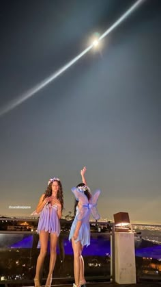 two beautiful women standing on top of a roof at night with an airplane in the sky