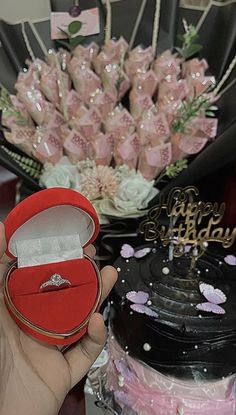 a person holding a ring box in front of a birthday cake and bouquet of flowers