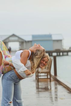 two women are hugging on the dock in front of some water and buildings, one is holding her back