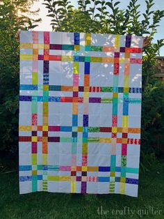 a colorful quilt hanging on a clothes line in front of some bushes and trees with the sun behind it