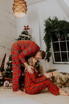 a man and woman dressed in red plaid pajamas kissing while sitting on the floor next to a christmas tree