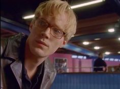 a man with glasses and a leather jacket looks at the camera while standing in a building