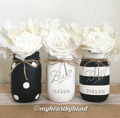 three painted mason jars with white roses in them on top of a burlie table