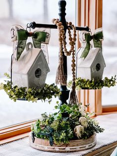 two birdhouses are hanging from a stand with flowers and greenery in front of a window