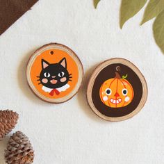 two wooden slices with cartoon pumpkins and cat faces on them, sitting next to pine cones