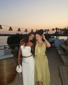 two women standing next to each other in front of a body of water at sunset