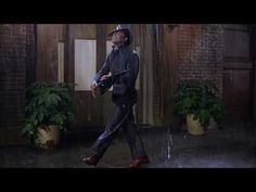 a man in a suit and hat walking through the rain with an umbrella over his head