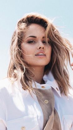 a woman with long hair standing in front of a blue sky wearing a trench coat