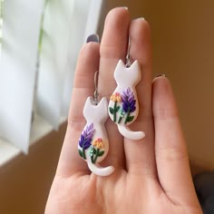 a person holding two cat shaped earrings in their hand with flowers painted on the back