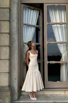 a woman standing in front of a window wearing a white dress