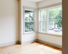 an empty room with two windows and hard wood flooring on one side, looking out onto the street