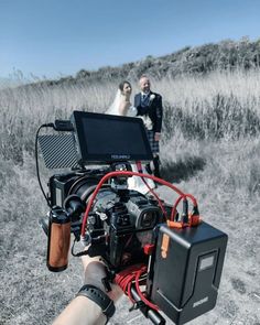 a person holding a video camera in front of a bride and groom on a hill