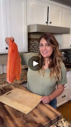 a woman standing in a kitchen holding up a piece of fish on a cutting board