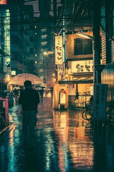 a man with an umbrella walks down the street in the rain at night, while buildings are lit up behind him