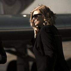 a woman in sunglasses is walking near an airplane and talking on her cell phone as she looks off to the side