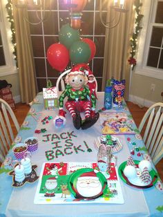 a table set for christmas with presents and balloons in the background, including an elf doll