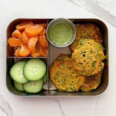 a metal container filled with food and cucumber next to orange slices on a table