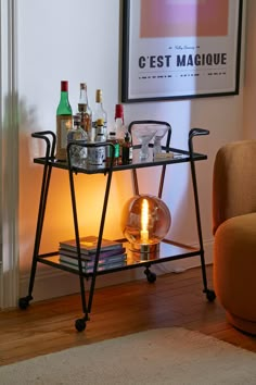 a bar cart with drinks on it in front of a framed poster and orange chair