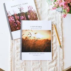 two spiral notebooks sitting on top of a table next to a pen and flowers