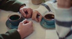 two people sitting at a table with cups of coffee in front of them and one person writing on a piece of paper