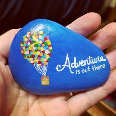 a hand holding a blue painted rock that says adventure is out there with a hot air balloon on it