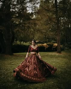 a woman in a red and gold dress standing in the grass with trees behind her