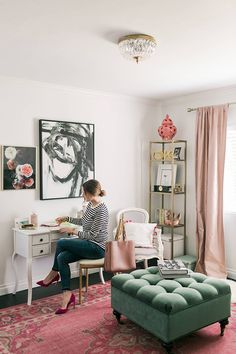 a woman sitting at a desk in front of a painting on the wall next to a green ottoman