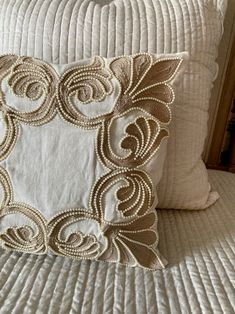 a close up of a pillow on a bed with white linens and gold threadwork