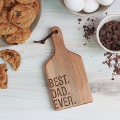 a wooden cutting board with the words best dad ever on it next to chocolate chip cookies and eggs