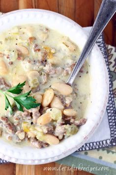 a white bowl filled with beans and meat soup on top of a wooden table next to a spoon