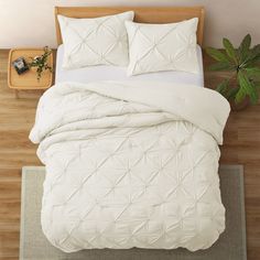 a bed with white comforter and pillows on top of it next to a plant